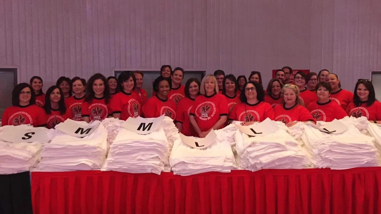 Group of people wearing matching red t-shirts standing behind large stacks of white t-shirts sorted by size